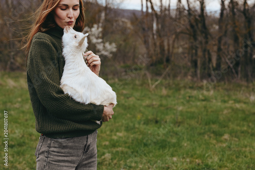 Foto A woman in a knitted sweater cradles a white goat kid outdoors in a grassy field, sunlight filtering through trees