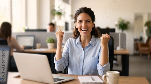 Joyful woman celebrates success at her desk. She grips her fists, eyes gleaming with triumph. Laptop rests open before her, notebook beside it. Cafe-style office hums with soft daylight and greenery