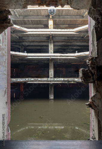 a multi-tiered concrete cavern stands in quiet desolation, viewed through a jagged, crumbling breach in a foreground wall. The rough, broken edges of a severely damaged wall frame the view, acting