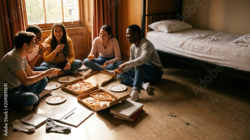 Young friends eating pizza on dorm room floor with notebooks nearby casual college life laughter shared meal student community weekend break affordable housing social connection youth culture