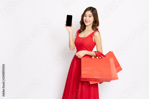 Happy Asian woman in a red dress holding red shopping bags and showing a blank smartphone screen, ideal for e-commerce, mobile application, online sale, and retail business advertisement