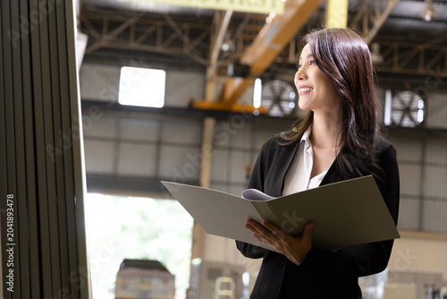 Successful Asian female manager in a black suit holding a folder and looking away in a warehouse, ideal for industrial business advertisement, logistics promotion, and factory marketing banner