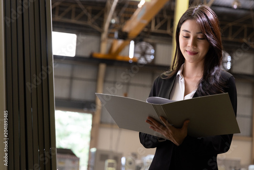 Successful Asian female manager in a black suit checking documents in a folder inside a warehouse, ideal for industrial business advertisement, logistics promotion, and corporate factory marketing