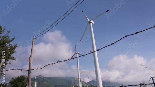 Wind turbines generating clean energy under a blue sky, partially obscured by power lines and barbed wire