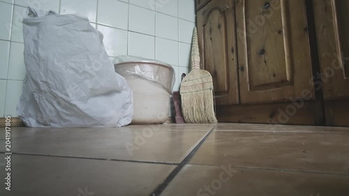 Still life scene of a domestic kitchen corner. Low angle shot showing a tiled floor with a paper sack, a bucket and a straw broom