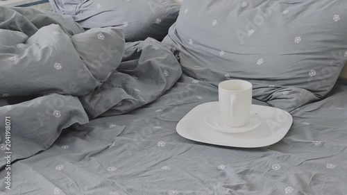 Messy gray bed with wrinkled sheets and pillows. An empty white coffee cup sits on a plate, suggesting a peaceful morning spent in bed
