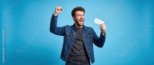 Excited young man celebrating big win holding ticket on blue background