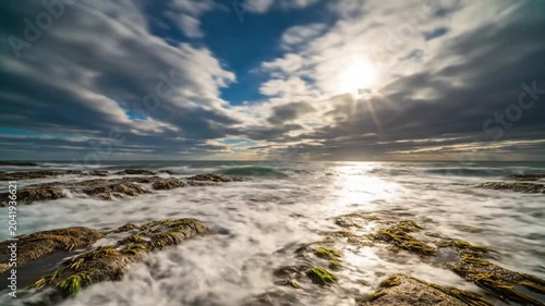 Time-lapse of ocean waves crashing against coastal rocks under a dynamic cloudy sky, sunlight shining