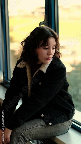 Upset teenage girl sits curled on a windowsill indoors, avoiding eye contact and thinking about problems at school and social media, concept emotional burnout and stress in youth.
