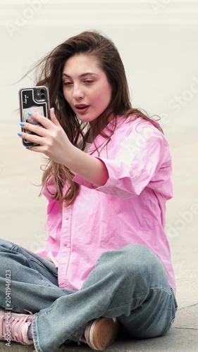Casual urban girl with long hair sits on the ground and lifts smartphone toward the camera, posing for front camera shot and creating fun content for a short video app.