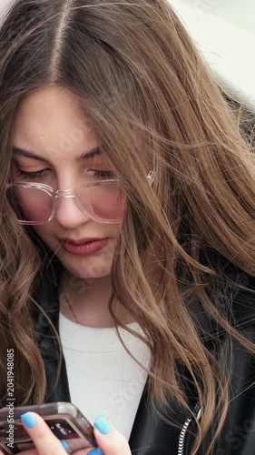 Anxious young woman with pink tinted glasses stares at her phone, silently reading a stressful message and processing unexpected news.