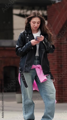 Vertical video of a stylish young woman in leather jacket sitting down and then standing up again, smiling at the camera and playfully engaging with the viewer in a windy urban setting.