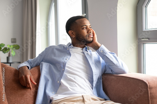 Bild auf Leinwand A young, mature African-American man sitting on the couch at home looking out the window daydreaming