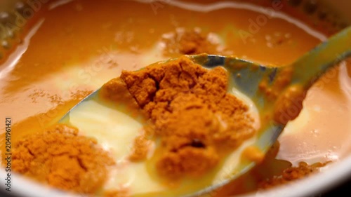Metal spoon lifting undissolved turmeric powder particles from rice beverage during Ayurvedic golden milk medicine preparation