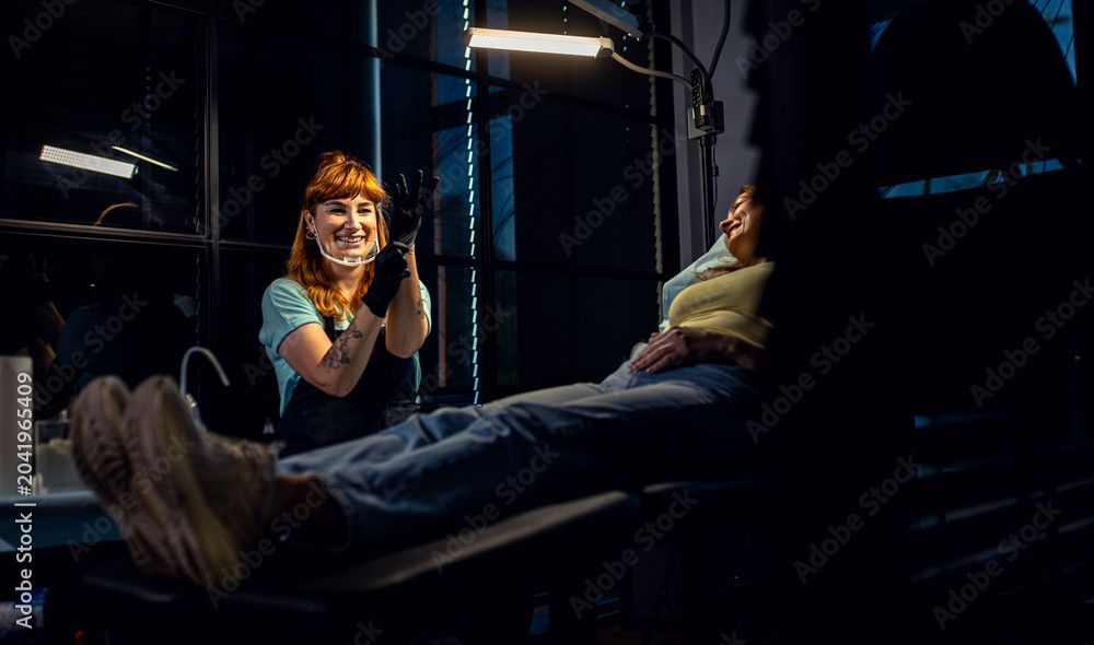 Fototapeta premium Female tattoo artist in a modern studio puts on gloves and prepares to make a tattoo on a female client.