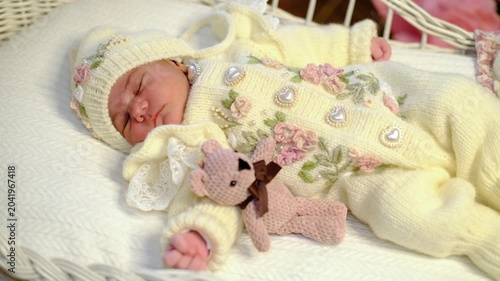 A gentle side view frames a newborn asleep in a white wicker cradle, wearing floral knitwear beside a tiny teddy toy. Use it for first days, baby care, cozy nursery style, and tender family stories.