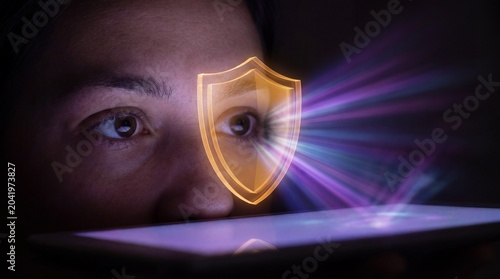 Close-up of a person looking at a tablet with a glowing shield symbol over their eye, representing online security and protection in a dark setting with vibrant light rays.