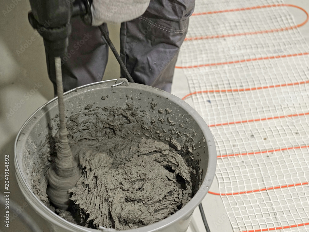 Obraz premium Construction worker preparing tile surface, mixing mortar substrate for flooring, underfloor heating mat. Workman combines adhesive in renovation setting. Operating power tool during bathroom upgrade