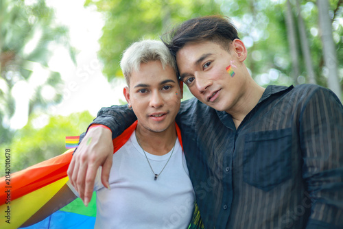 Gay couple LGBTQ community strolled through nature park waving brightly colored Prime Minister's flag openly displaying affection through intimate hugs kisses demonstrating proud connection community.