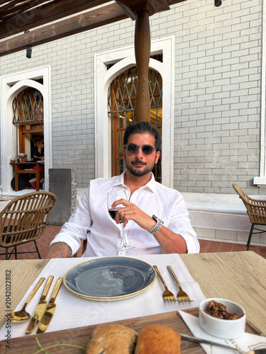 Handsome dark-haired man in a linen shirt sitting at a local Greek island restaurant, holding a glass of wine, elegant pose, sunset ambiance, seaside dining, Mediterranean lifestyle