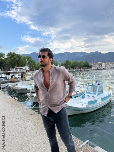 Handsome dark-haired man in a linen shirt on a Greek island at sunset, small boats in the background, calm sea, golden hour, Mediterranean lifestyle