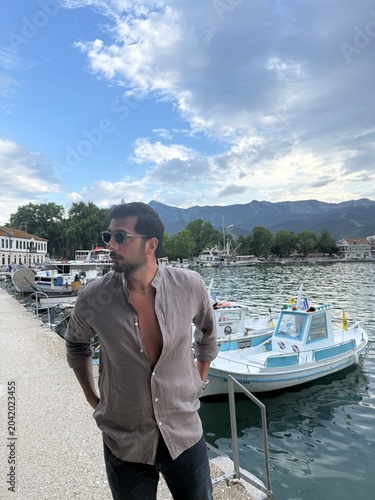 Handsome dark-haired man in a linen shirt on a Greek island at sunset, small boats in the background, calm sea, golden hour, Mediterranean lifestyle