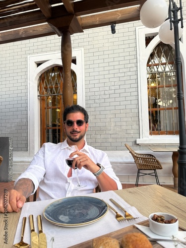 Handsome dark-haired man in a linen shirt sitting at a local Greek island restaurant, holding a glass of wine, elegant pose, sunset ambiance, seaside dining, Mediterranean lifestyle