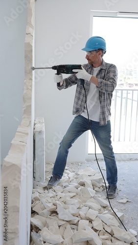 Male construction worker wearing grey checked shirt and blue hardhat is demolishing a wall with a hammer drill, creating construction debris on the surrounding floor surface. Renovation concept