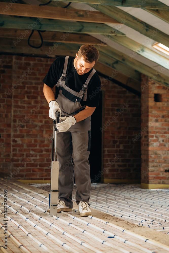 Fototapeta premium Male construction worker in gray overalls using a tool to install flooring in a rustic attic with exposed brick walls and wooden beams overhead