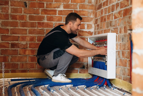 Foto Male plumber in black shirt and gray overalls adjusts heating system manifold on