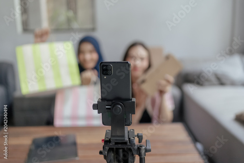 A smartphone mounted on a tripod records two blurred people holding colorful shopping bags in a bright living room with a sofa and coffee table in the background