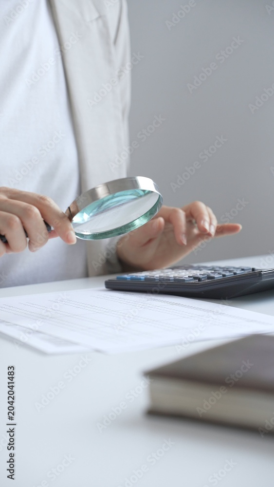 Fototapeta premium Businesswoman carefully reviewing financial reports using magnifying glass and calculator, close up vertical view. Audit and taxes in business