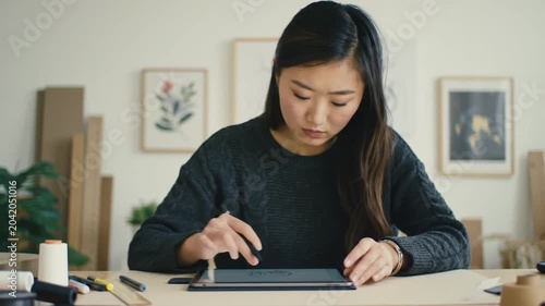 Asian Woman Designs on Tablet at Desk with Art.