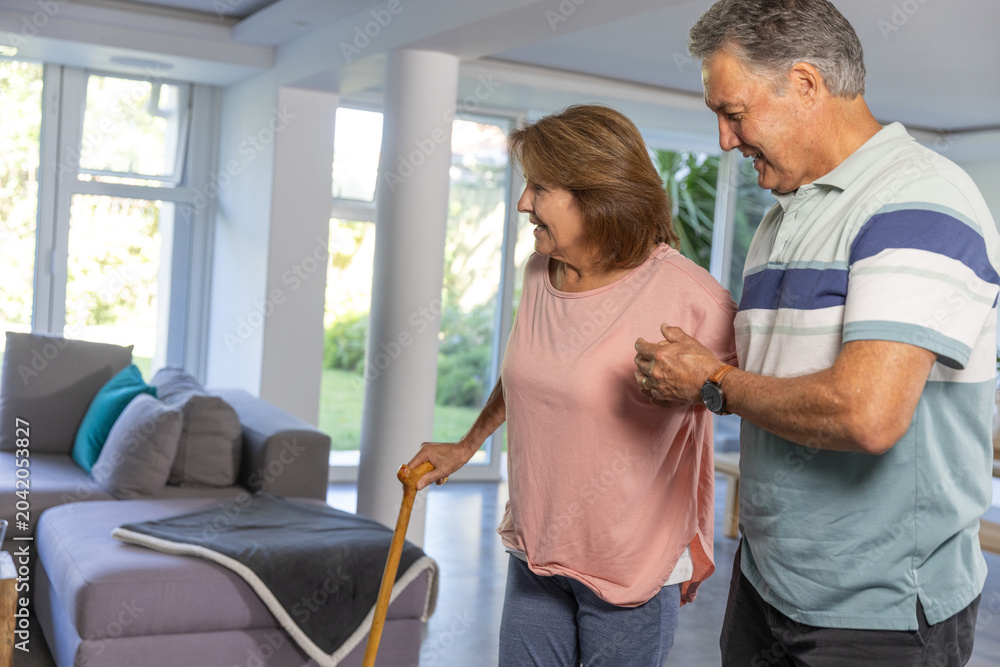 Naklejka premium Mature man in striped polo steadying Senior woman in top holding wooden cane in living room