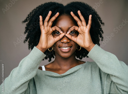 Close up portrait of attractive quirky young black woman making binoculars with hands showing ok gesture on white studio background