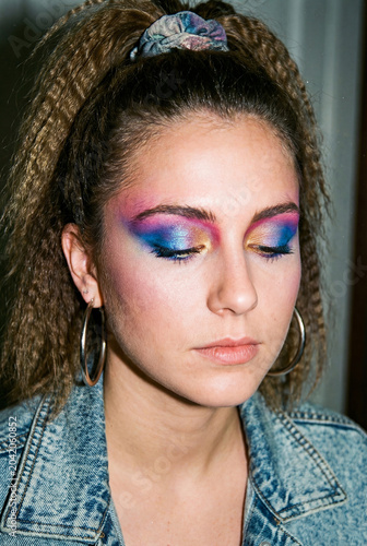A portrait of a woman with multicolored eyeshadow and 80s-style makeup, wearing a denim jacket