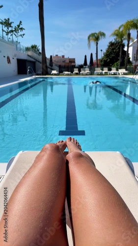 pov of woman legs on sun lounger with man swimming laps in resort pool, authentic summer vacation lifestyle