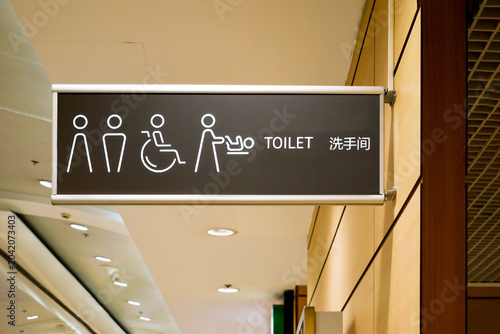 Suspended black signboard with white icons for male, female, disabled access, and diaper changing station, featuring English and Chinese text in a public building interior space area.