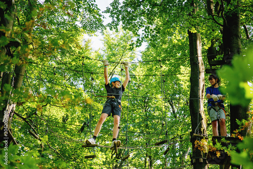 Two teenager boys climbing rope course in forest adventure park, teenagers wearing helmets and harnesses enjoying outdoor challenge, real life. Brothers and best friends outdoors.