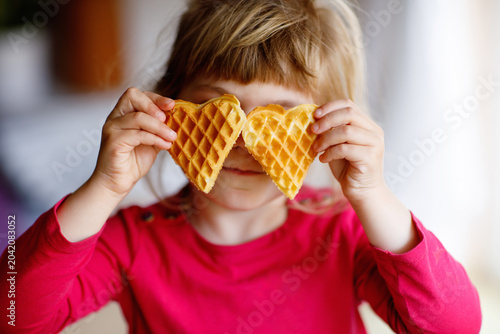 Portrait of happy little preschool girl holding fresh baked heart waffle. Smiling hungry toddler child with sweet biscuit wafer. Sweet sugar belgian waffles.