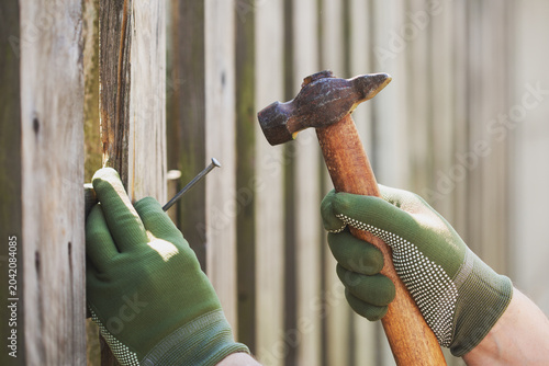 Male worker hands drive nail with hammer tool in rusrtic old wooden fence plank, maintenance repair, diy and carpentry outdoor.