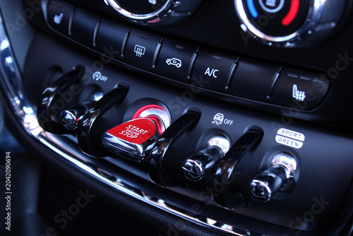 Сar dashboard featuring a glowing red engine start stop button, shiny chrome rotary knobs, and various control switches. This sleek compact design utilizes high-grade glossy plastic and metallic eleme