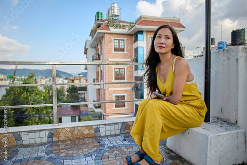 Female model in mustard dress sitting on rooftop ledge legs crossed gazing into distance