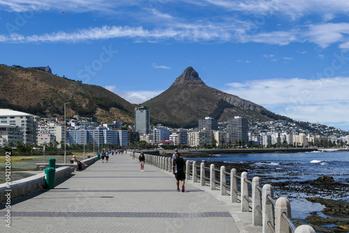 People walk along the Sea Point promenade on a sunny day in Cape Town, with Lion's Head in the distance