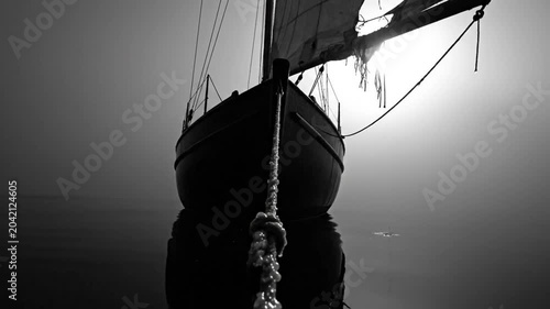 Black and white shot of a classic sailboat at anchor with the sun illuminating the sail and reflecting in the still water