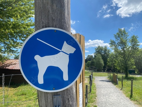 Hinweisschild für angeleinte Hunde entlang eines Spazierwegs im Grünen. Symbol für Hundehaltung, Leinenpflicht, Rücksichtnahme und Regeln in öffentlichen Bereichen. Echtfoto.