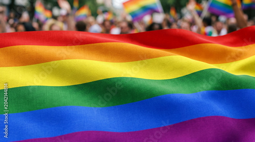 Rainbow flag waving in close up with blurred crowd of people holding flags during lgbtq pride parade, symbol of lgbt community, love, freedom and diversity