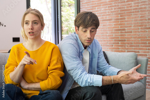 Fotografie Young couple arguing on a sofa in a modern living room, woman pointing while man looks frustrated; domestic relationship conflict, tense conversation and emotional disagreement