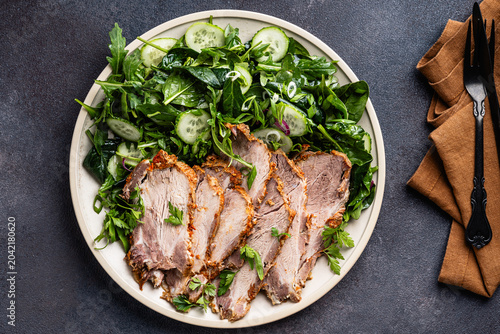 Slices of pork meat roast served with green vegetables salad in plate, top view
