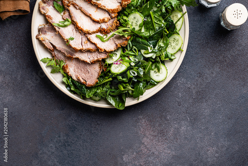 Slices of pork meat roast served with green vegetables salad in plate, top view